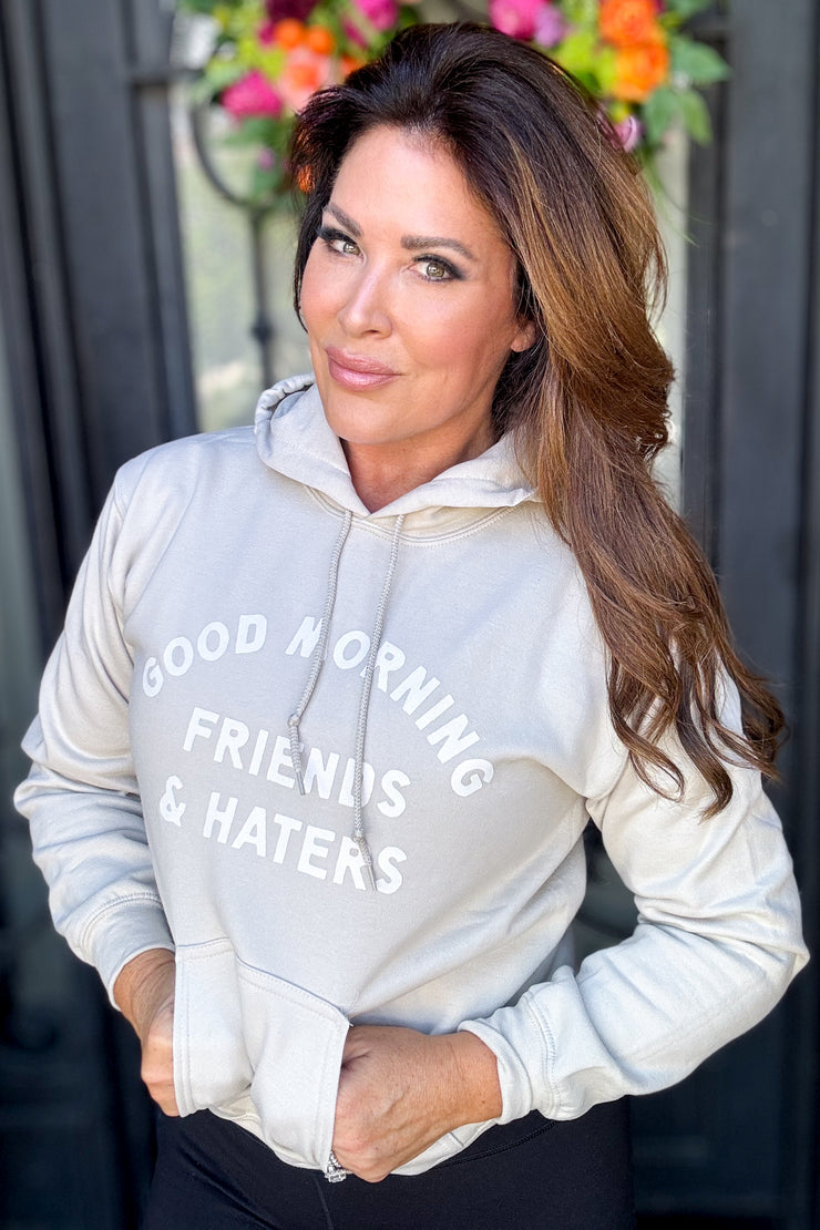 Woman wearing a tan hooded sweatshirt with "Good Morning Friends & Haters" printed on middle front of sweatshirt in white writing. 