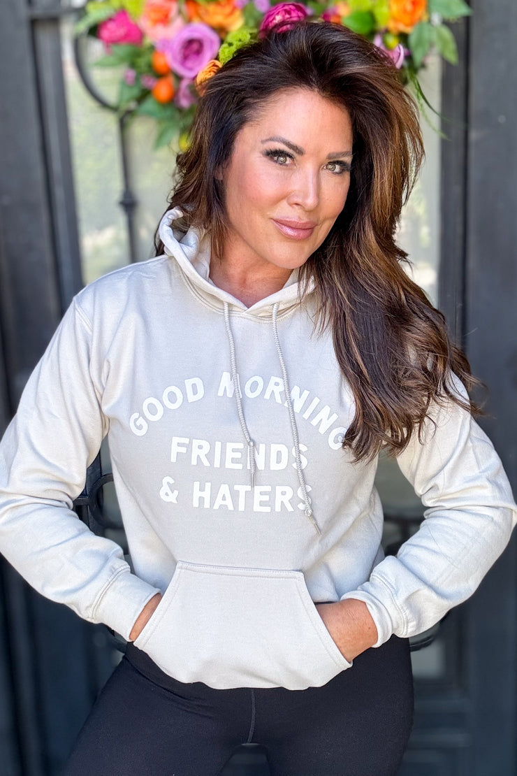 Woman wearing a tan hooded sweatshirt with "Good Morning Friends & Haters" printed on middle front of sweatshirt in white writing. 