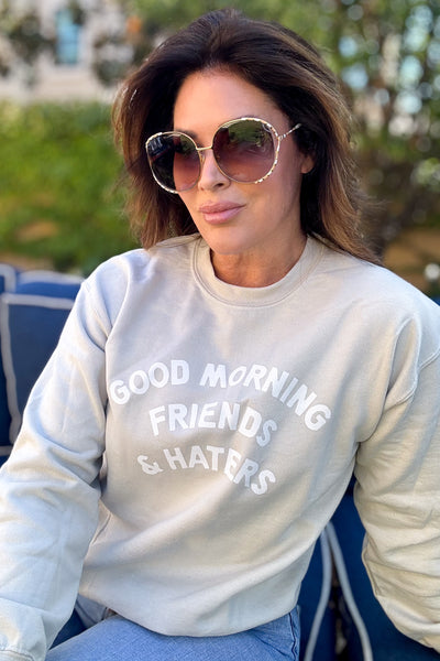 Woman in a tan crewneck sweatshirt  that says good morning friends and haters