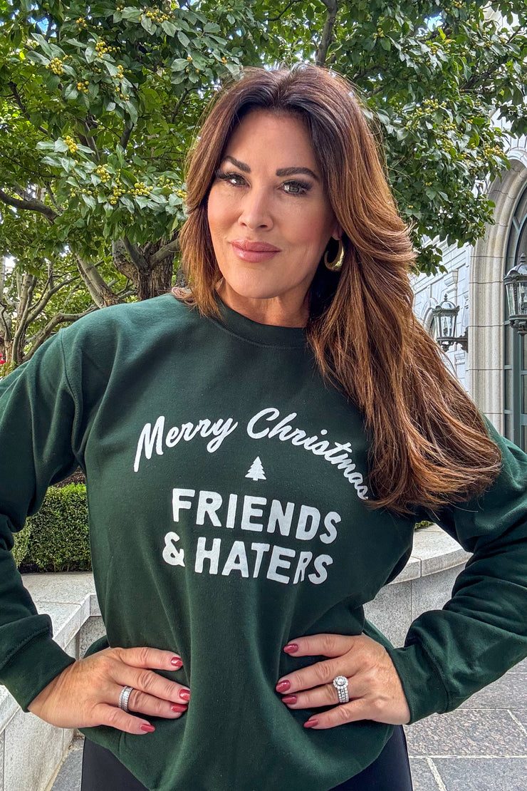 Woman wearing a green crewneck sweatshirt with "Merry Christmas Friends & Haters" printed on middle front of sweatshirt in white writing. 